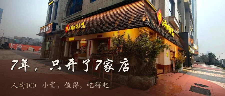 实拍封面：田小狗饭店沿街门面，仿小青瓦屋檐、夯土色墙、门口竹丛与陶罐、脚下红砖地，黄底手写体招牌，门边立着写“立春”的节气木牌，头顶是现代写字楼的灰色石材幕墙