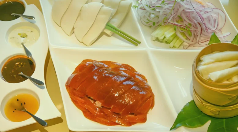 一只片好的北京烤鸭铺在盘里，配荷叶饼、甜面酱、葱白与黄瓜条，枣红油亮